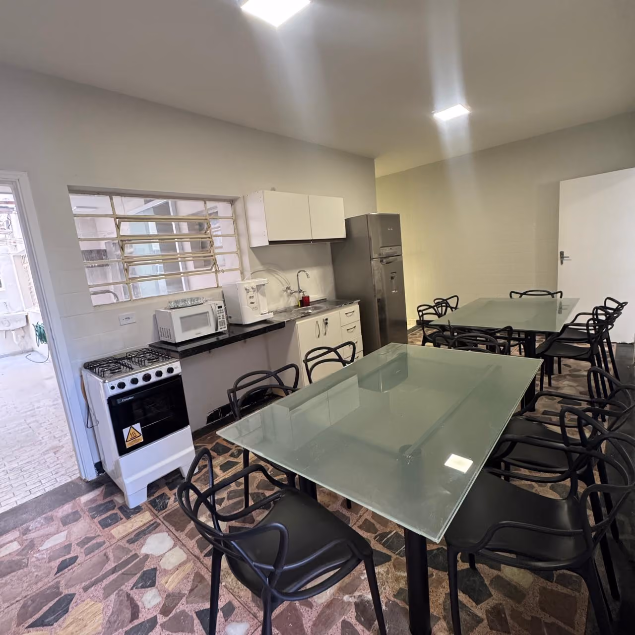 Cozinha pequena com bancada de granito clara que tem cafeteira, fogão elétrico de duas bocas, pia de inox com escorredor, micro-ondas e frigobar branco, parede com grande espelho e porta de madeira com aviso de proibido fumar.