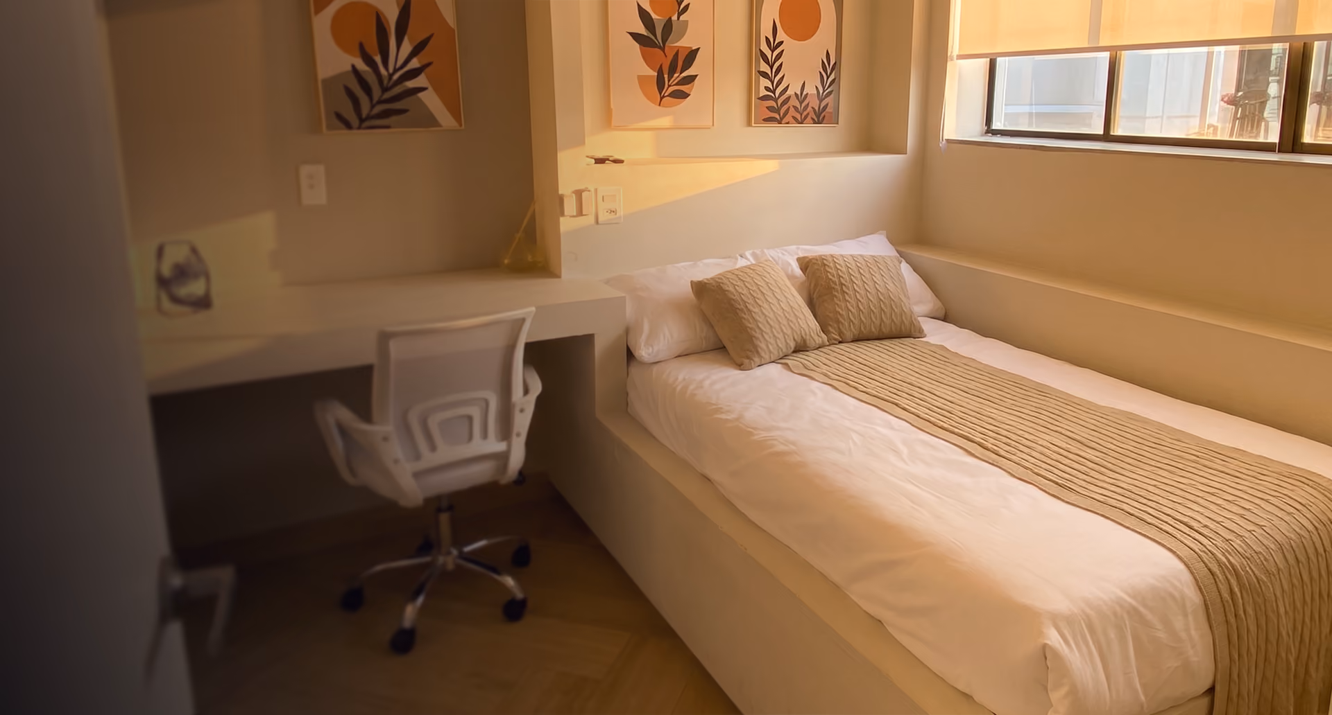 Quarto minimalista com cama de solteiro coberta por edredom branco, jogos de almofadas e uma manta bege trançada, mesa de trabalho branca ao lado com cadeira giratória branca, janela grande com persiana e três quadros decorativos com arte botânica em tons de marrom e bege na parede.