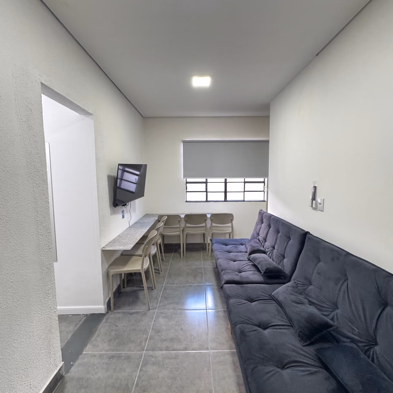 Quarto com duas beliches de estrutura metálica preta, cada uma com colchão branco, travesseiro e roupa de cama preta e branca, com duas pequenas mesas de madeira presas às camas, chão de ladrilhos claros e paredes com pintura azul e verde, com janela e aparelho de ar-condicionado instalado na parede.