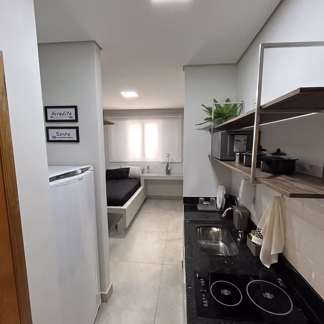 Apartamento pequeno com cozinha compacta à direita, incluindo pia, fogão elétrico e prateleiras com utensílios; ao fundo, cama de solteiro com almofadas, mesa de trabalho sob a janela com cortina branca; parede à esquerda com geladeira e quadros escritos 'Acredite' e 'Sonhe'.