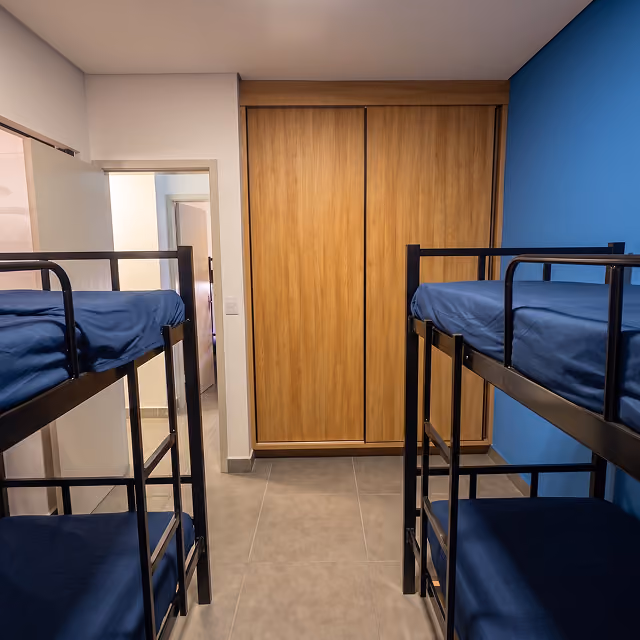 Quarto com duas beliches pretas com colchões e lençóis azuis, parede azul à direita e armário embutido de madeira ao fundo.
