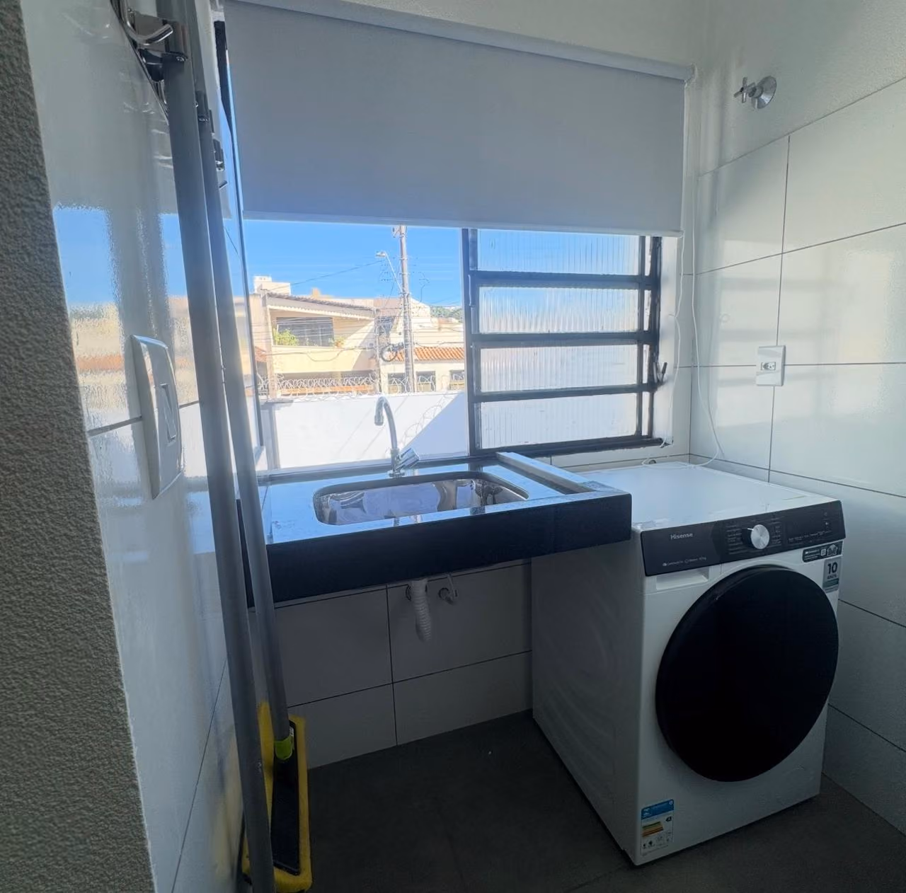 Cozinha pequena com bancada de granito clara que tem cafeteira, fogão elétrico de duas bocas, pia de inox com escorredor, micro-ondas e frigobar branco, parede com grande espelho e porta de madeira com aviso de proibido fumar.