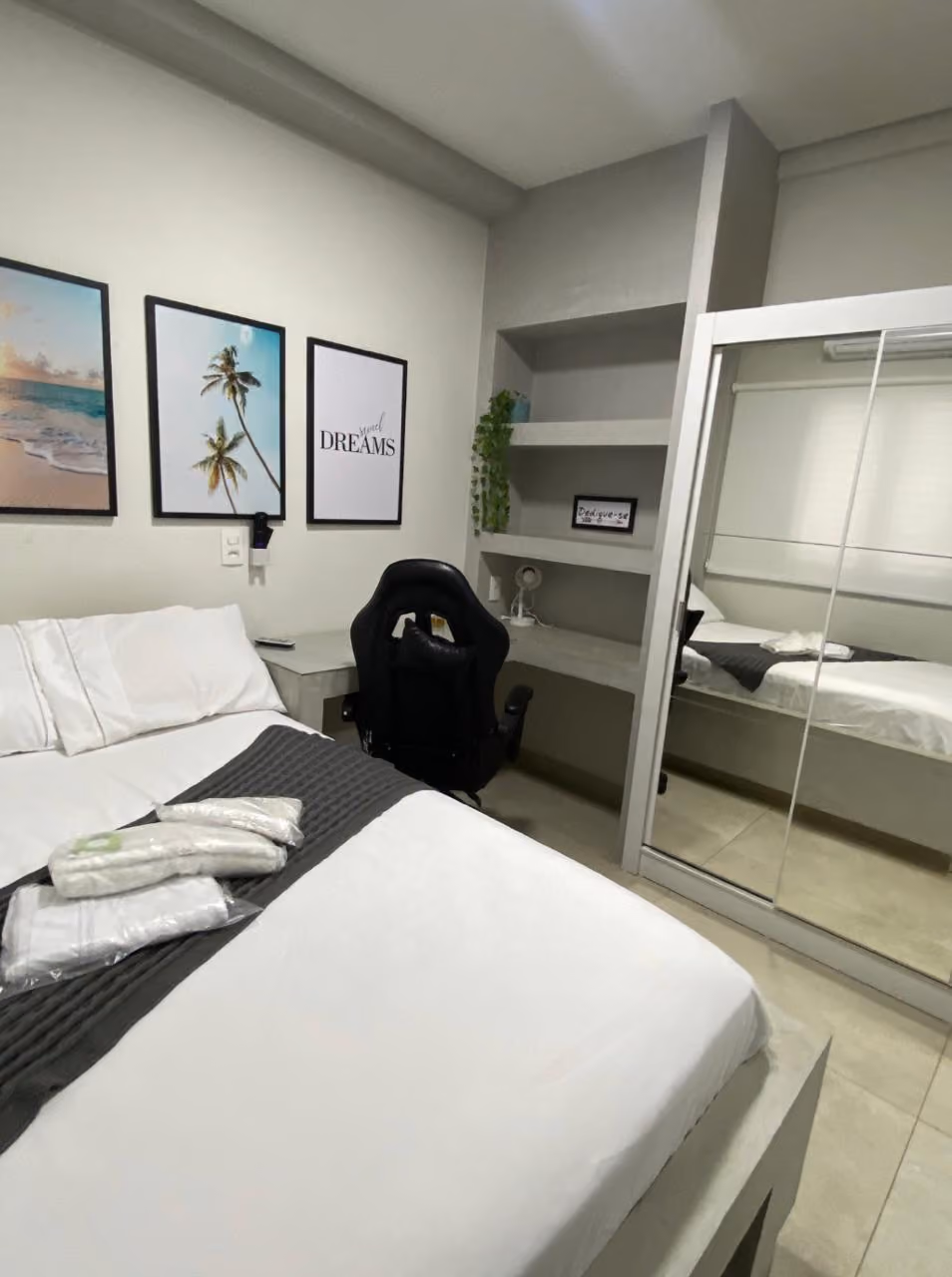 Sala de espera iluminada com três poltronas cinza dispostas em fila ao lado de paredes claras com textura ondulada, pequenas mesas redondas brancas entre as cadeiras, um capacho e uma planta alta verde perto da porta branca ao fundo.